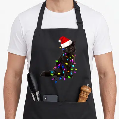 Tablier de cuisine noir avec un chat noir portant un bonnet rouge et des guirlandes lumineuses colorées enroulées autour du corps.