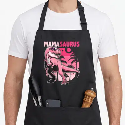 Tablier de cuisine noir avec un T Rex rose aux lunettes de soleil et le texte Mamasaurus en arrière-plan tropical.
