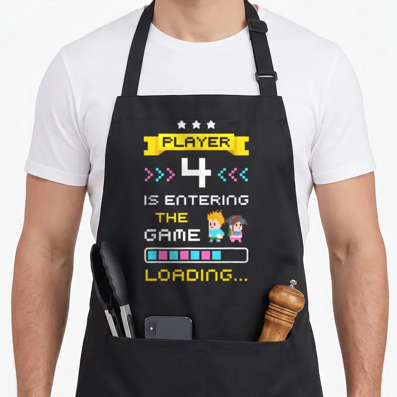 Tablier de cuisine noir avec illustration pixelisée Player 4 is entering the Game Loading et deux personnages colorés.