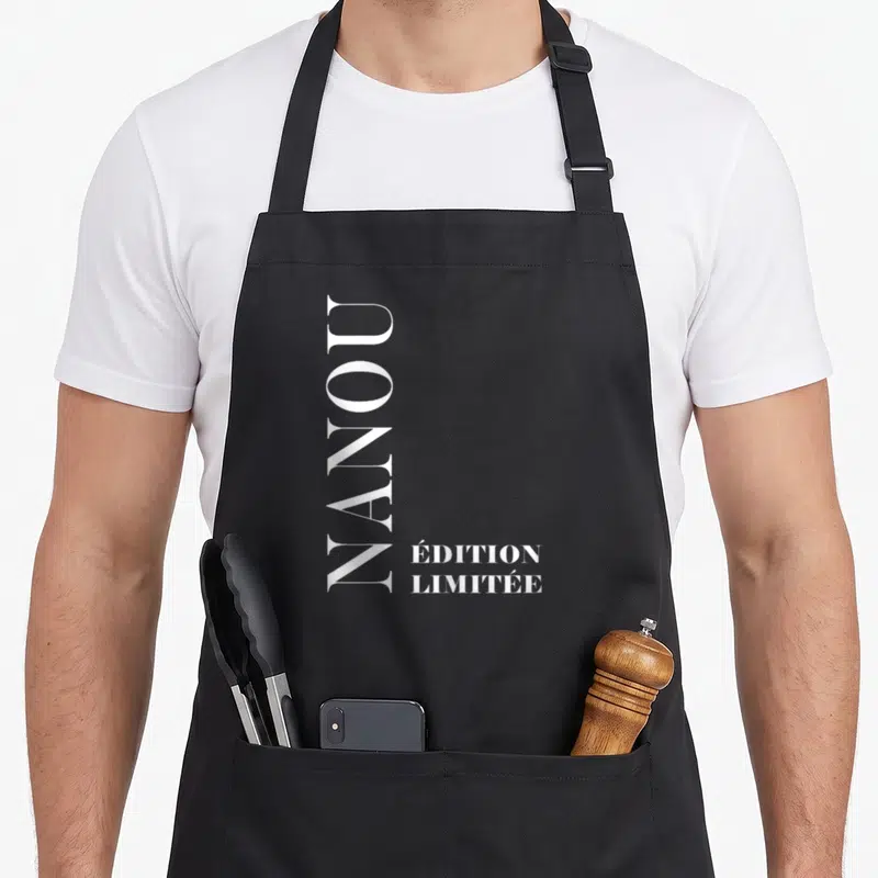 Tablier de cuisine noir avec l'inscription « Nanou » en lettres blanches verticales sur le devant.