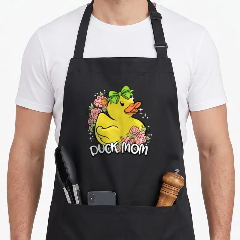 Tablier de cuisine noir avec illustration canard jaune coiffé d'un nœud vert entouré de fleurs roses et du texte DUCK MOM