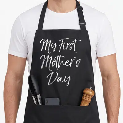 Tablier de cuisine noir avec l'inscription blanche My First Mother's Day en écriture cursive élégante.