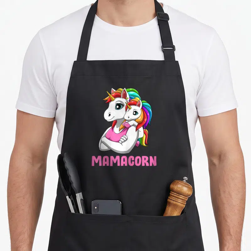 Tablier de cuisine noir avec illustration de deux licornes blanches s'enlaçant et du texte MAMACORN en rose.
