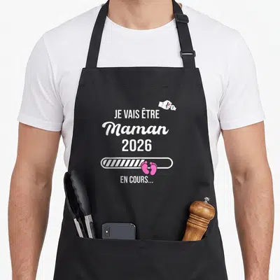 Tablier de cuisine noir avec le texte Je vais être Maman 2026 et une barre de chargement rose ornée de petits pieds.