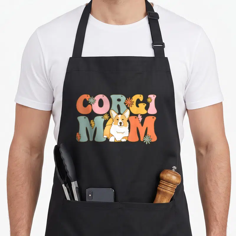 Tablier de cuisine noir avec illustration Corgi Mom Groovy en lettres pastel et chien souriant au centre du design.