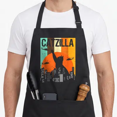 Tablier de cuisine noir avec illustration rétro d'un chat géant Catzilla sur fond de ville et soleil orange.