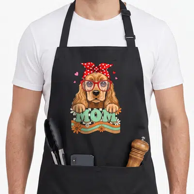 Tablier de cuisine noir avec un cocker épagneul portant des lunettes rouges et le mot MOM en lettres vertes.