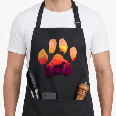 Tablier de cuisine noir avec une grande patte de chien colorée et une silhouette de labrador au milieu d'un paysage forestier.