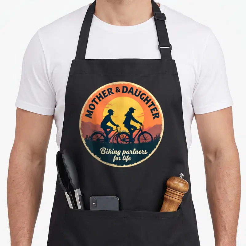Tablier de cuisine noir avec un motif circulaire orange représentant deux cyclistes en silhouette sous le soleil.