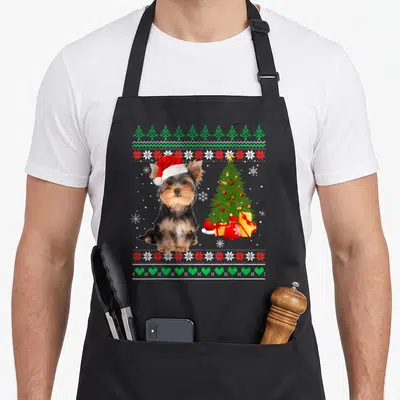 Tablier de cuisine noir avec un chien Yorkshire portant un bonnet de Noël près d'un sapin décoré et de cadeaux.