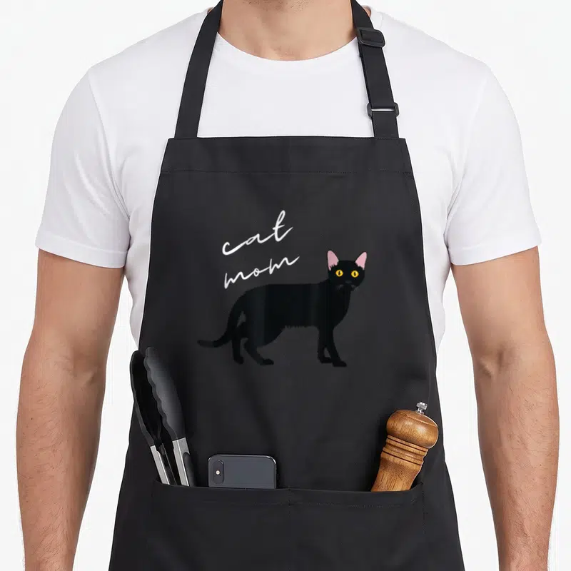 Tablier de cuisine noir avec illustration d'un chat noir aux yeux jaunes et texte cat mom en blanc.