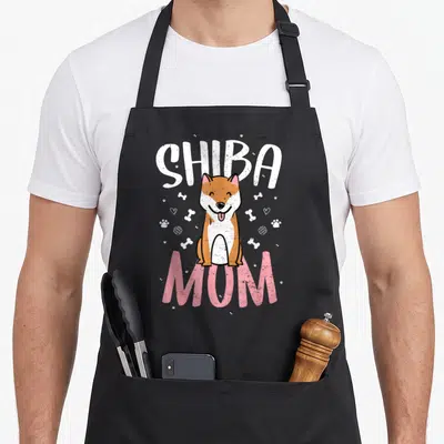 Tablier de cuisine noir avec illustration d'un Shiba Inu souriant et les mots Shiba Mom en lettres blanches et roses.