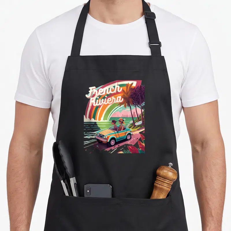 Tablier de cuisine noir avec illustration rétro French Riviera montrant une voiture sur la côte méditerranéenne.