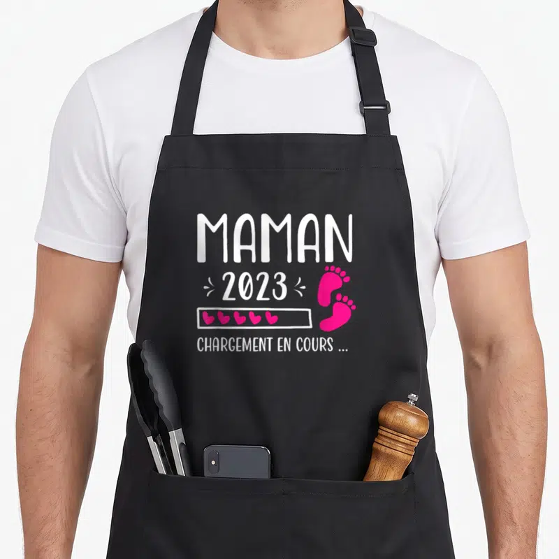 Tablier de cuisine noir avec l'inscription Maman 2023 Chargement en cours et des empreintes de pieds roses.