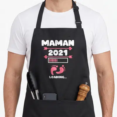 Tablier de cuisine noir avec texte Maman 2021 Loading et petits pieds roses stylisés.