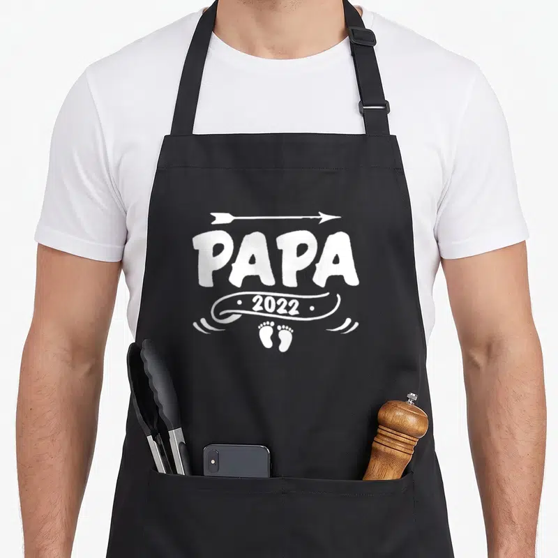 Tablier de cuisine noir avec inscription Papa 2022, flèche blanche et empreintes de petits pieds en bas du motif central.