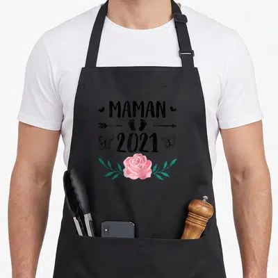 Tablier de cuisine noir avec texte Maman 2021, empreintes de pieds, papillons et une rose rose en bas du motif imprimé.