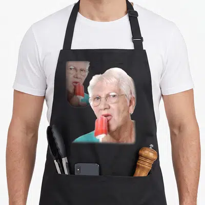 Tablier de cuisine noir avec une illustration d'une femme âgée portant des lunettes et mangeant un sorbet rouge sur bâtonnet.