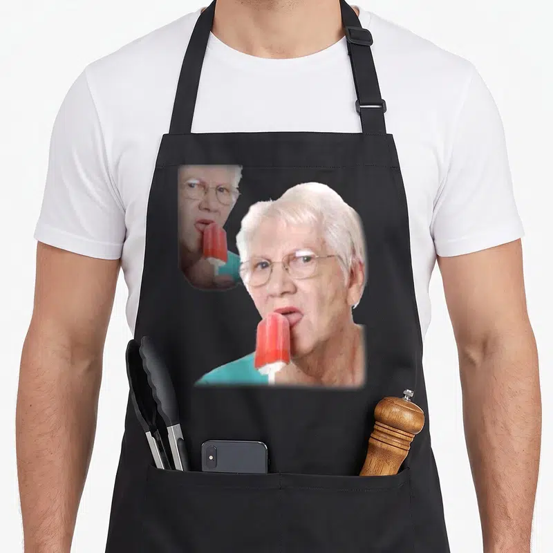 Tablier de cuisine noir avec une illustration d'une femme âgée portant des lunettes et mangeant un sorbet rouge sur bâtonnet.