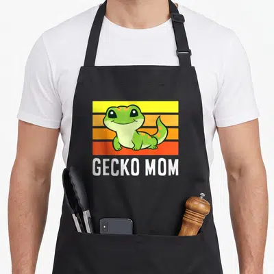 Tablier de cuisine noir avec un gecko vert souriant sur fond de bandes jaunes oranges et rouges et l'inscription GECKO MOM.