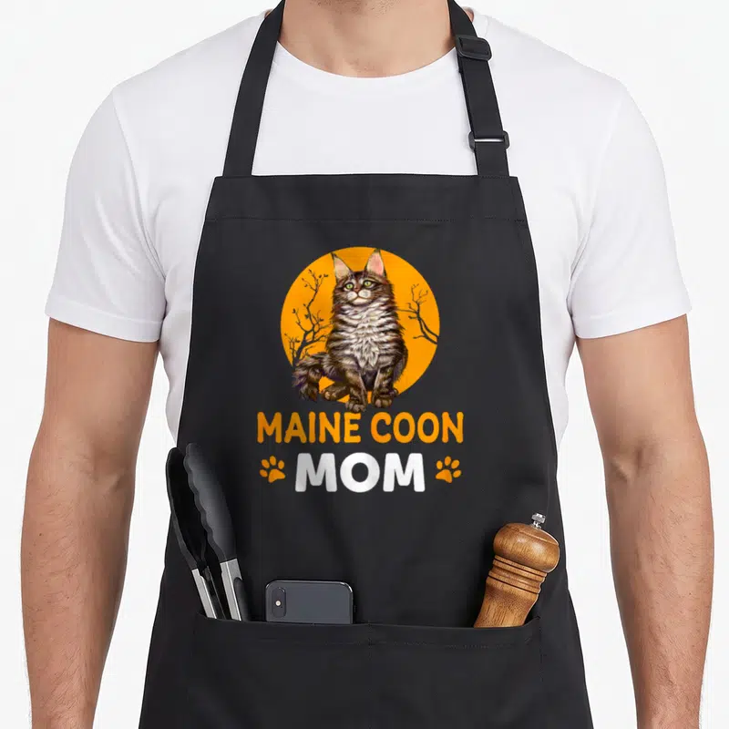 Tablier de cuisine noir avec illustration Maine Coon Mom et empreintes de pattes oranges sur fond circulaire.