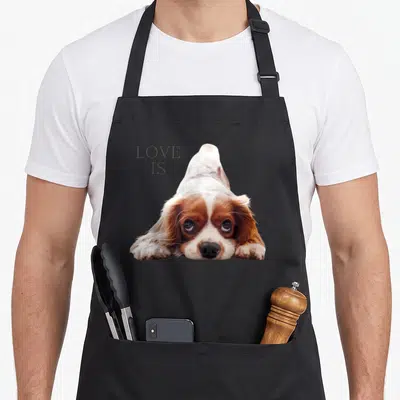 Tablier de cuisine noir avec un chien Cavalier King Charles Spaniel et le texte Love Is imprimé en gris.