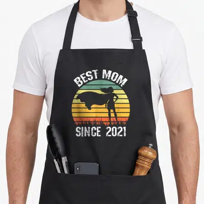Tablier de cuisine noir avec silhouette de super-maman et texte Best Mom Since 2021 sur fond de soleil rayé.