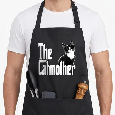 Tablier de cuisine noir avec le texte The Catmother et un chat noir et blanc regardant vers le haut.