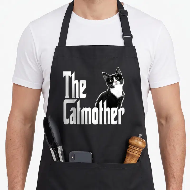 Tablier de cuisine noir avec le texte The Catmother et un chat noir et blanc regardant vers le haut.