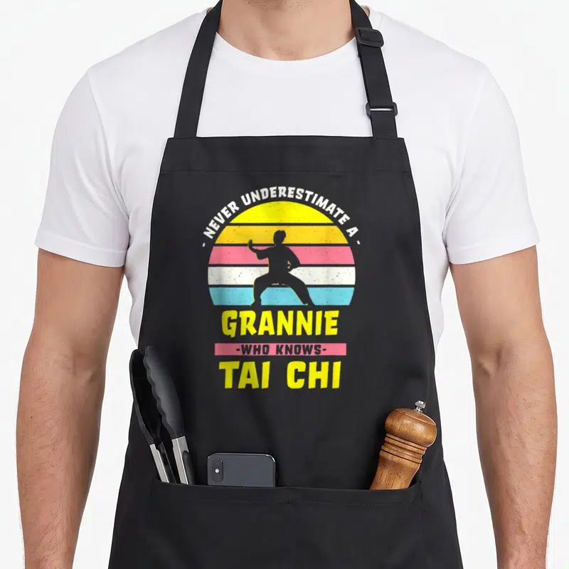 Tablier de cuisine noir avec une silhouette de grand-mère en posture de tai chi devant un soleil arc-en-ciel vintage.