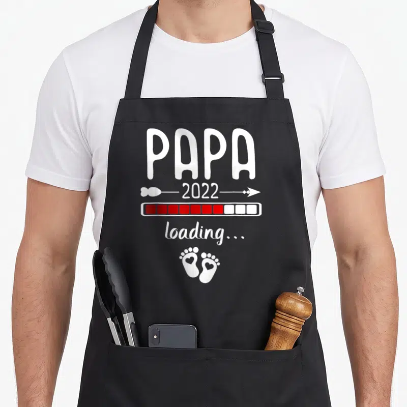 Tablier de cuisine noir avec inscription Papa 2022 loading et petits pieds blancs ornés d'un cœur.