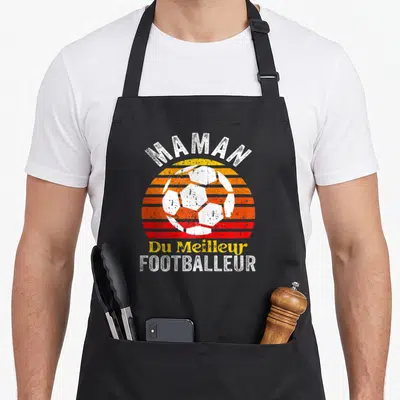 Tablier de cuisine noir avec un ballon de foot blanc sur un soleil orange et rouge, le mot Maman en haut et Footballeur en bas.