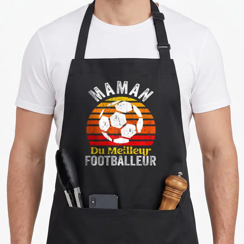Tablier de cuisine noir avec un ballon de foot blanc sur un soleil orange et rouge, le mot Maman en haut et Footballeur en bas.