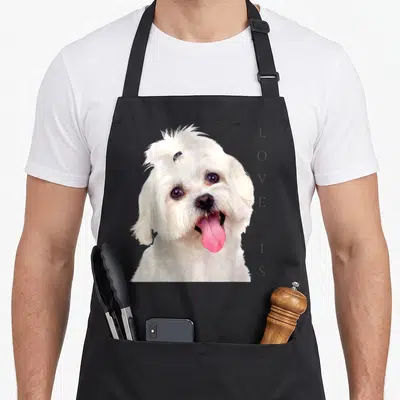Tablier de cuisine noir avec portrait d'un bichon maltais blanc et texte vertical love is sur le côté droit.