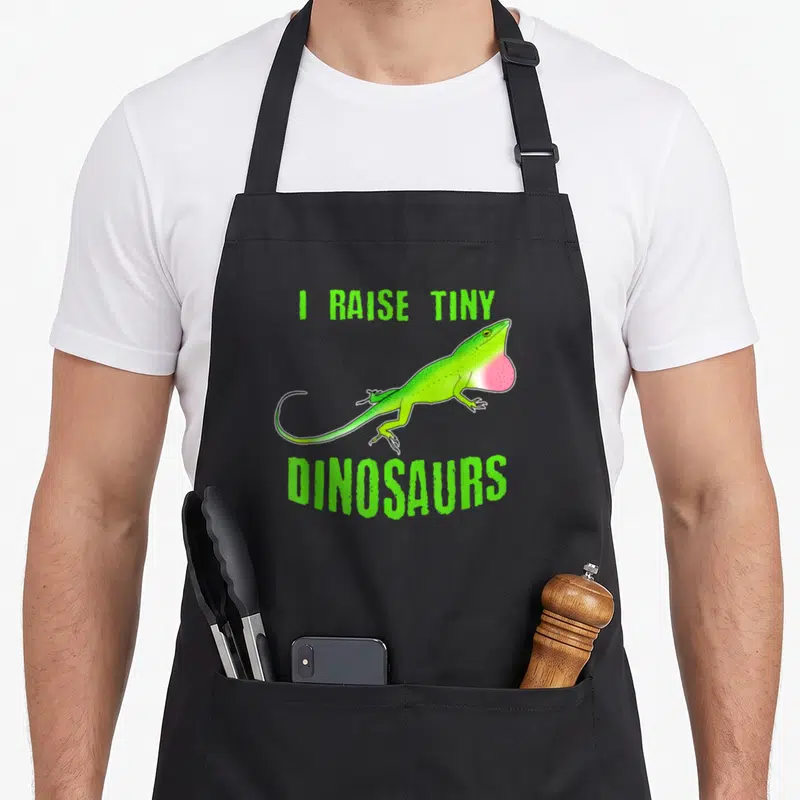 Tablier de cuisine noir avec un lézard anole vert et l'inscription I raise tiny dinosaurs en lettres vertes.