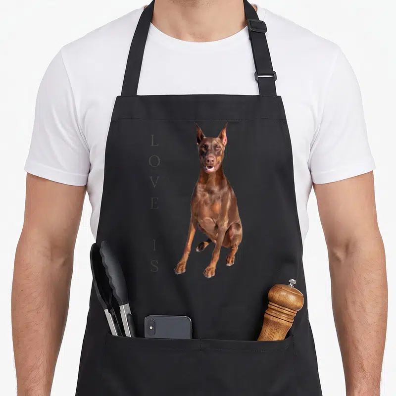 Tablier de cuisine noir avec un Doberman brun assis et le texte Love Is vertical à côté.