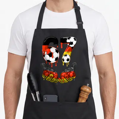 Tablier de cuisine noir avec le chiffre 67 aux couleurs allemande, des ballons de football et deux mains faisant un signe positif.