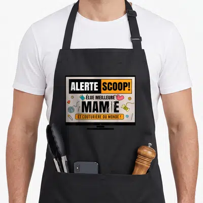 Tablier de cuisine noir avec illustration télévision affichant alerte scoop élue meilleure mamie et couturière du monde entourée d'outils de couture colorés.
