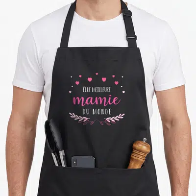Tablier de cuisine noir avec inscription Élue Meilleure Mamie du Monde entourée de cœurs roses et de feuillage stylisé.