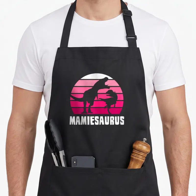 Tablier de cuisine noir avec illustration de deux dinosaures gris devant un soleil rose strié et le mot Mamiesaurus en bas.