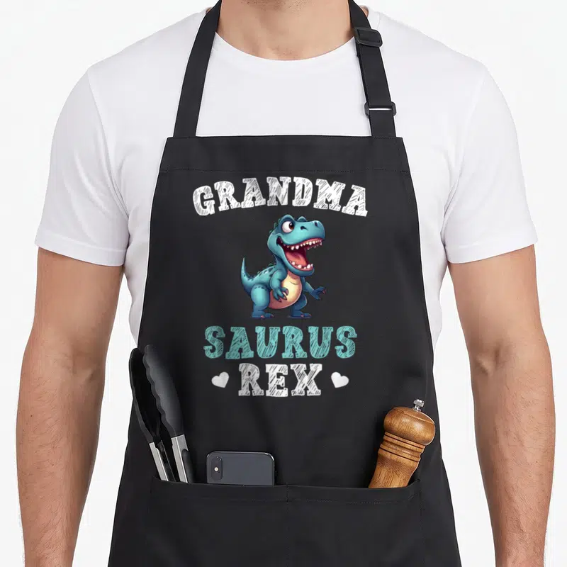 Tablier de cuisine noir avec un dinosaure bleu stylisé et les mots Grandma Saurus Rex encadrés de cœurs blancs.