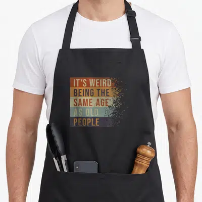 Tablier de cuisine noir avec inscription multicolore C'est bizarre d'avoir le même âge que de dire drôle vieux sur fond uni.