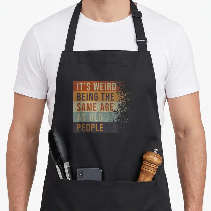 Tablier de cuisine noir avec inscription multicolore C'est bizarre d'avoir le même âge que de dire drôle vieux sur fond uni.