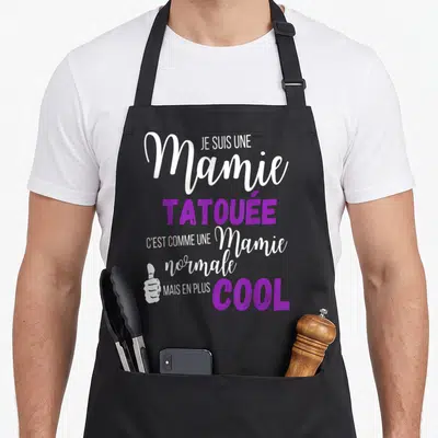 Tablier de cuisine noir avec texte blanc et violet affirmant être une mamie tatouée cool et un pouce levé stylisé.