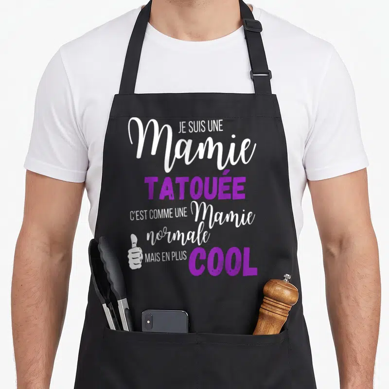 Tablier de cuisine noir avec texte blanc et violet affirmant être une mamie tatouée cool et un pouce levé stylisé.