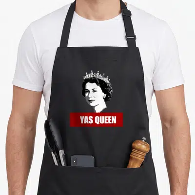 Tablier de cuisine noir avec portrait stylisé d'Elizabeth II et inscription Yas Queen sur bande rouge.