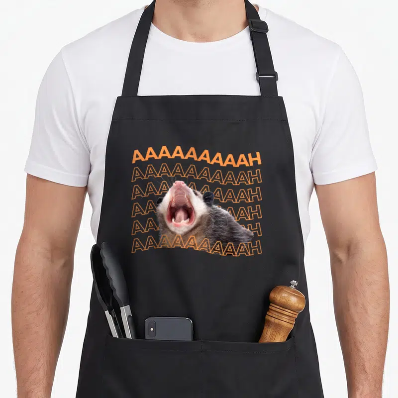 Tablier de cuisine noir avec un opossum hurlant et le texte AAAAAAAAAH en orange répété en arrière-plan.