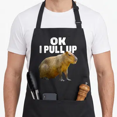 Tablier de cuisine noir avec un capybara et le texte blanc OK I PULL UP imprimé au centre.