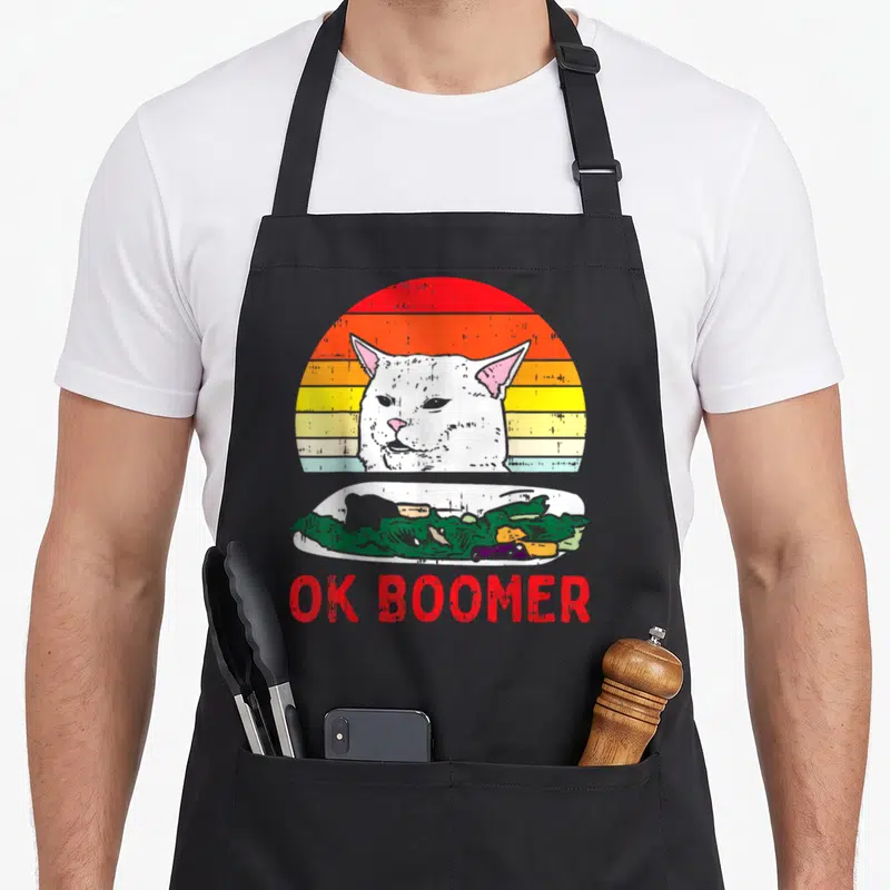 Tablier de cuisine noir avec chat blanc perplexe, plat vert et texte Ok Boomer rouge sur fond de soleil coloré.