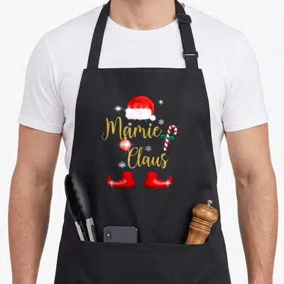 Tablier de cuisine noir avec chapeau de Père Noël, texte mamie Claus doré, canne à sucre et bottines rouges festives.